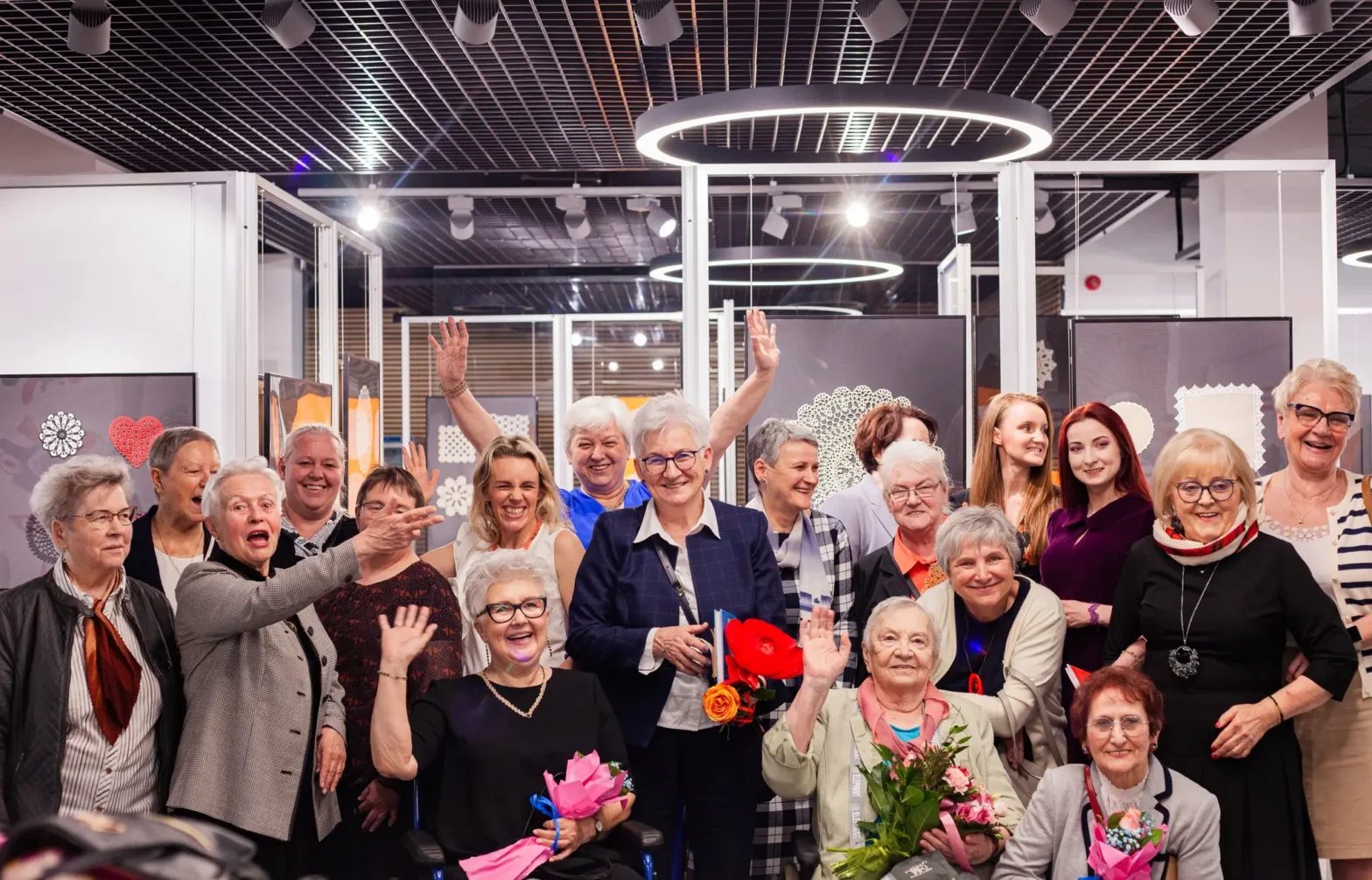 Grupa około dwudziestu uśmiechniętych kobiet w różnym wieku stoi i siedzi na tle wystawy z koronkowymi serwetkami w jasnym, nowoczesnym wnętrzu. Większość osób jest elegancko ubrana, kilka trzyma w rękach bukiety kwiatów. W centrum, na pierwszym planie, dwie kobiety siedzą na wózkach, trzymając bukiety. Wszyscy mają radosny wyraz twarzy, a niektóre osoby machają do obiektywu. W tle widać białe i kolorowe serwetki wyeksponowane na wystawie, co sugeruje, że grupa uczestniczy w wernisażu związanym z rękodziełem.