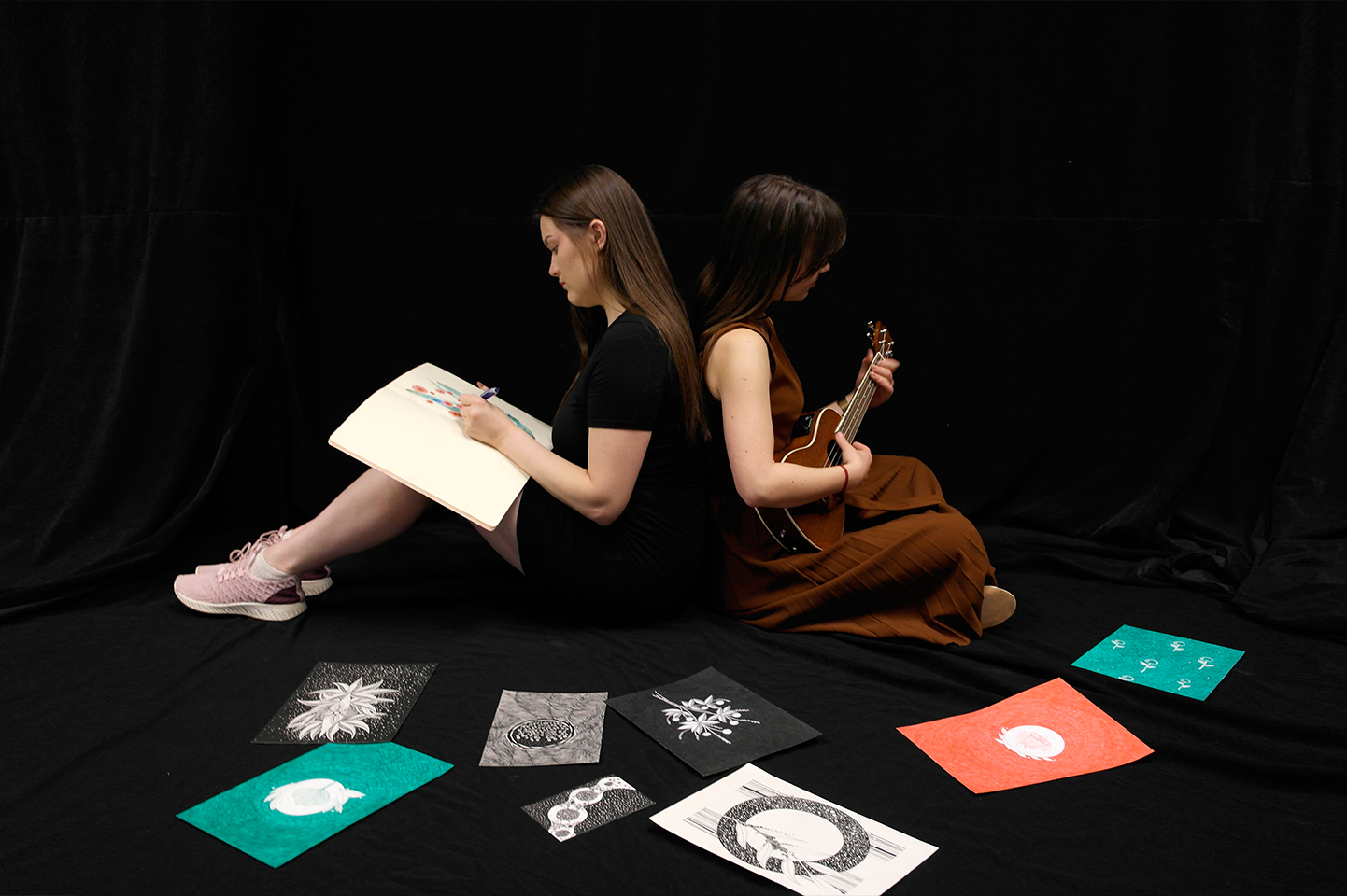 Dwie kobiety siedzą tyłem do siebie w czarnej przestrzeni. Jedna z nich ma szkicownik, w którym rysuje trzymając pisak lewą ręką, druga gran na ukulele. Przed nimi leżą ułożone w półokrąg rysunki.