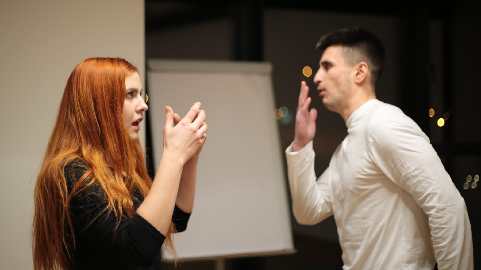 Scena teatralna. Młoda kobieta stoi z lewej strony i rozmawia z mężczyzną stojącym naprzeciwko gestykulując. Mężczyzna prawą dłonią zakrywa usta.