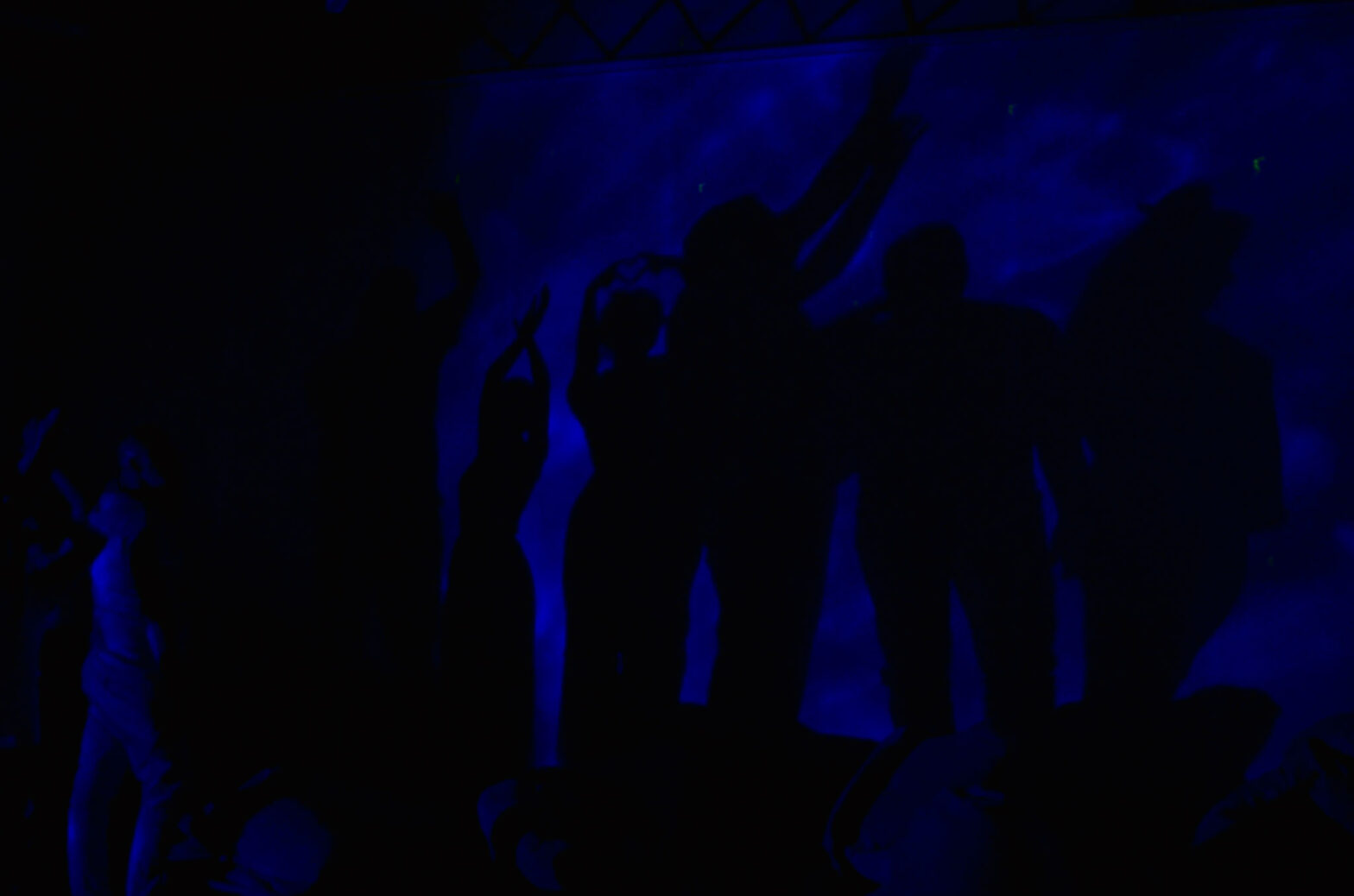 Grupa osób tańczy w ciemności. Oświetla je strumień niebieskiego światła. Światło pada na ścianę, na której powstają cienie tańczących. Każda osoba stoi w innej pozie. Jedna z osób składa dwa palce wskazujące i dwa kciuki w kształt serca.