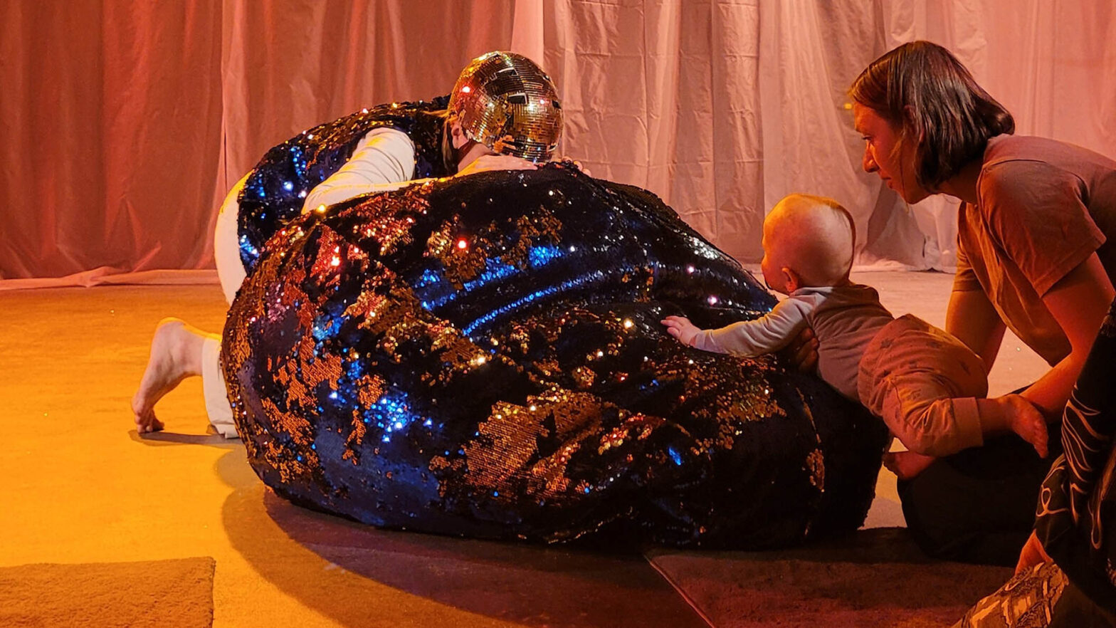 ALT: Spektakl Moja pierwsza odyseja kosmiczna. Aktorka w złotym hełmie ukrywa się za gigantyczną poduszką mieniącą się brokatem. Małe dziecko trzymane na rękach przez matkę przygląda się aktorce i wchodzi z nią w interakcję.