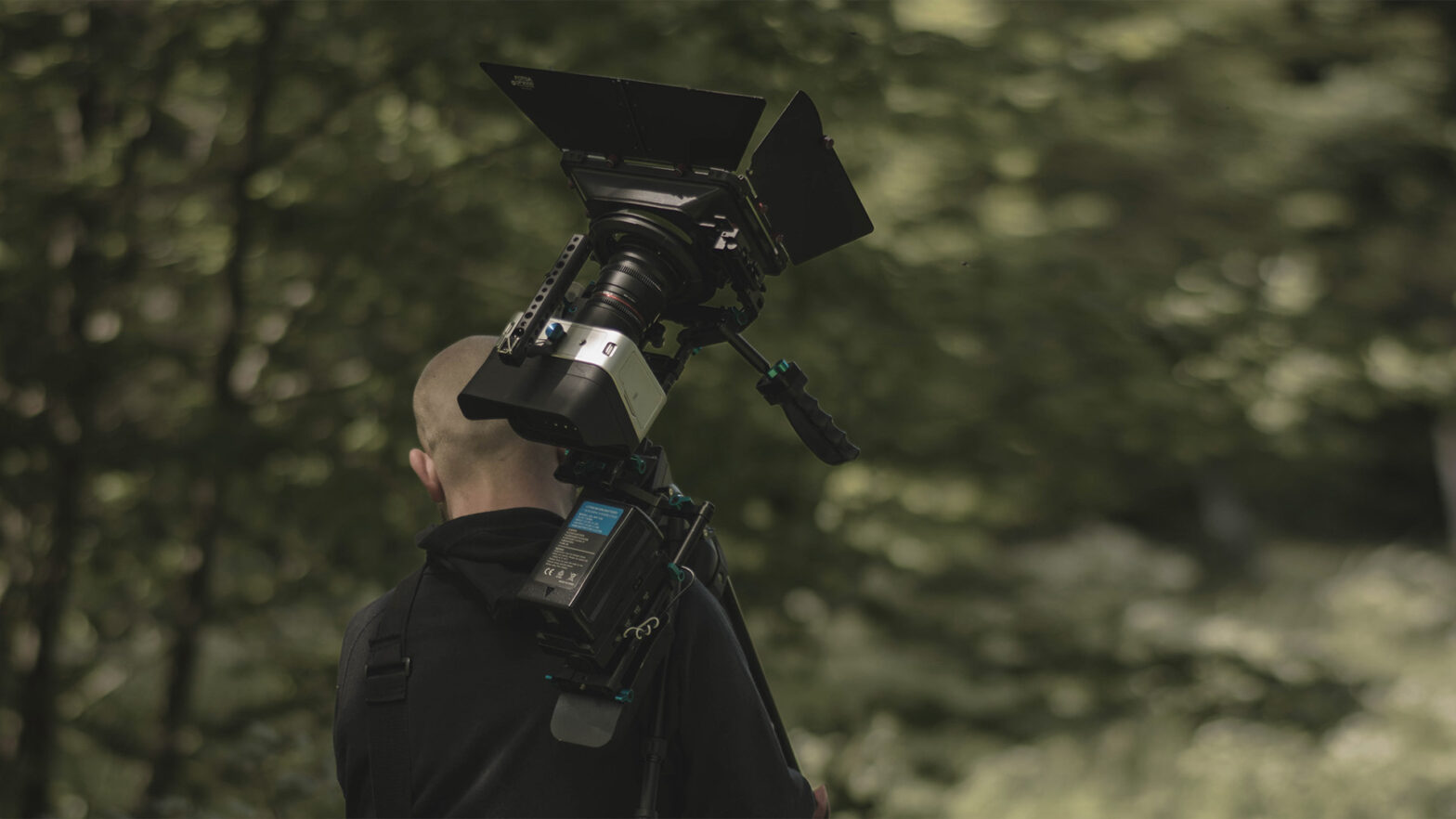 Pokazany od tyłu mężczyzna idzie przez las z kamerą filmową zamocowaną na statywie, który trzyma oparty o ramię.