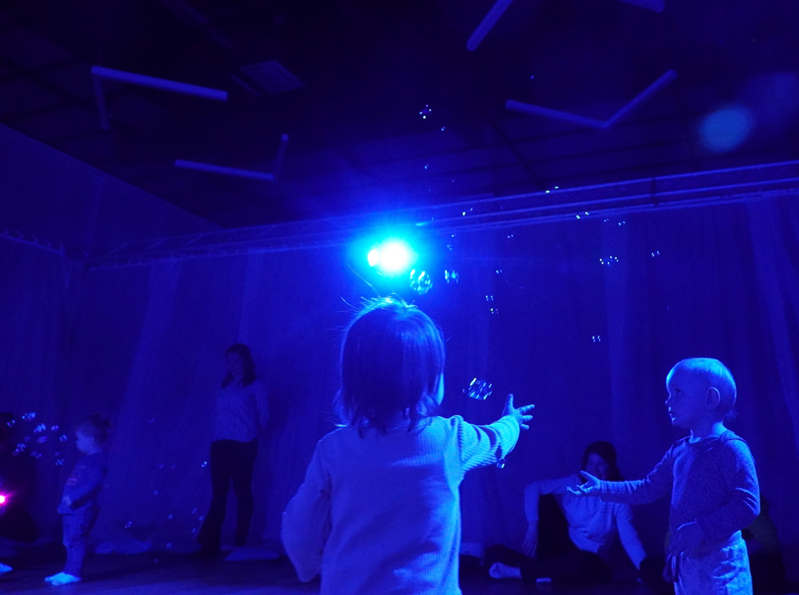 Małe dzieci bawią się bańkami mydlanymi w wyciemnionej przestrzeni, którą oświetla tylko jeden niebieski reflektor.
