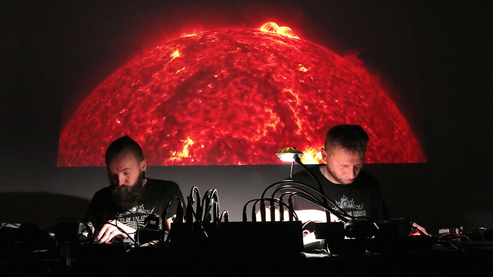 Muzycy podczas koncertu. Dwaj mężczyźni w skupieniu operują instrumentami elektronicznymi. W tle zdjęcie framgmentu Słońca z widocznym wyrzutem plazmy.