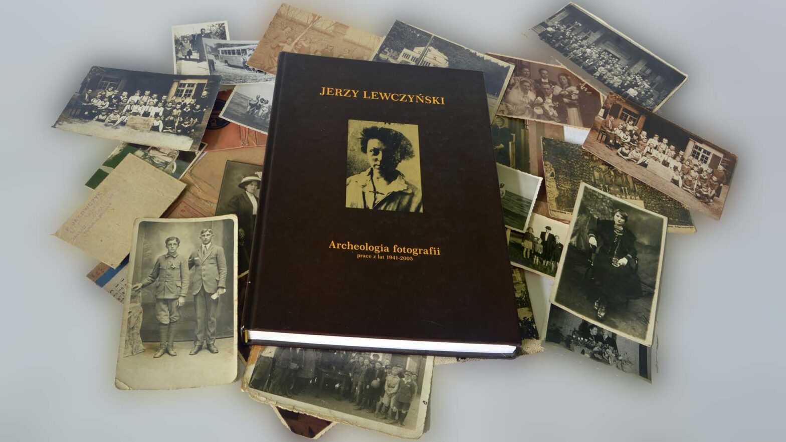 Stare zdjęcia rozrzucone na stole, na których leży książka Archeologia fotografii Jerzego Lewczyńskiego.