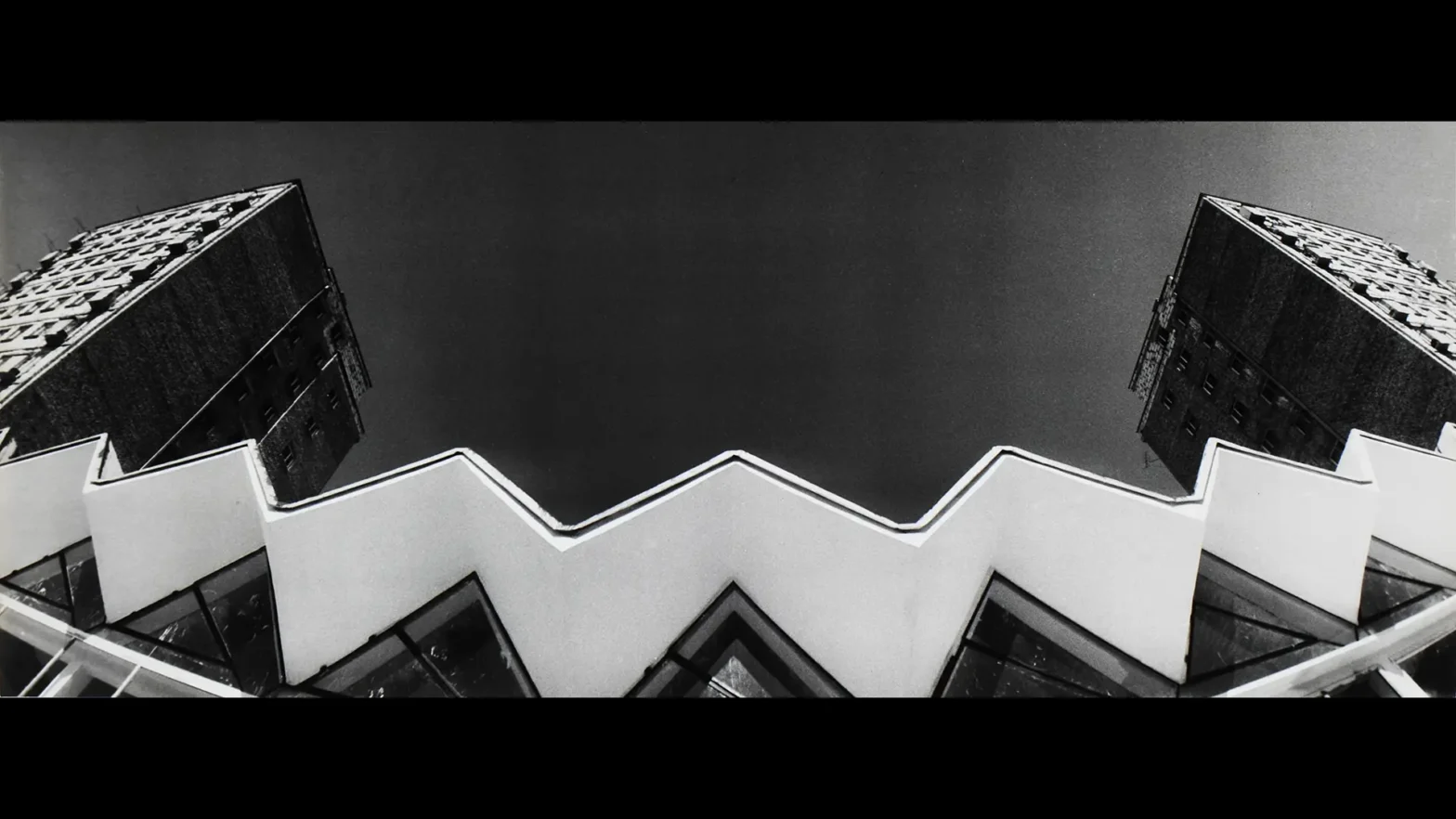 Autorem zdjęcia jest Ildefons Bańkowski, autorem reprodukcji zdjęcia jest Wojciech Woźniak. Zdjęcie przedstawia architekturę lat siedemdziesiątych. Wykonane jest z dołu w kolorze czarno białym. Przedstawia dwa bloki stojące na przeciwko siebie, połączone budowlą architektoniczną w kształcie zygzaka.