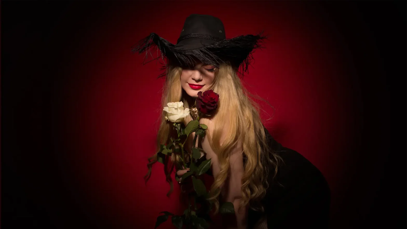 Na zdjęciu blond włosa kobieta. Na głowie ma czarny kapelusz, w ręku trzyma dwie róże. Jedna jest koloru białego, a druga czerwonego. Kobieta ma usta pomalowane czerwoną szminką i spuszczony wzrok w dół. Tło zdjęcia jest czerwone i delikatnie wyciemnione.