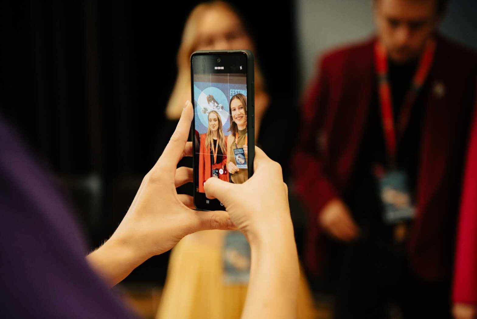 Na zdjęciu telefon trzymany w obu rękach. Osoba trzymająca telefon, robi nim zdjęcie. Na ekranie widoczne dwie kobiety, uśmiechające się do zdjęcia.