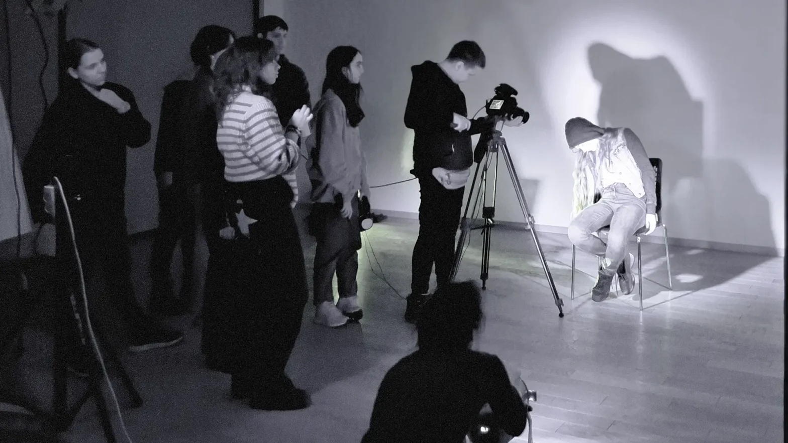 Czarno białe zdjęcie, na którym stoi grupa osób. Osoba stojąca na przodzie za aparatem, robi zdjęcie, osobie siedzącej przed obiektywem na krześle.