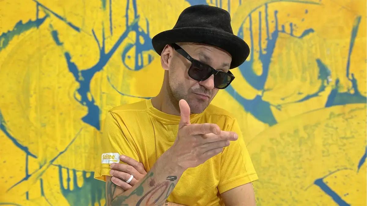 Na zdjęciu artysta Cebo, który na twarzy ma okulary, ubrany jest w czarny kapelusz i żółtą koszulkę. Za nim znajduje się na całej wielkości zdjęcia żółty obraz z błękitnymi śladami pędzla