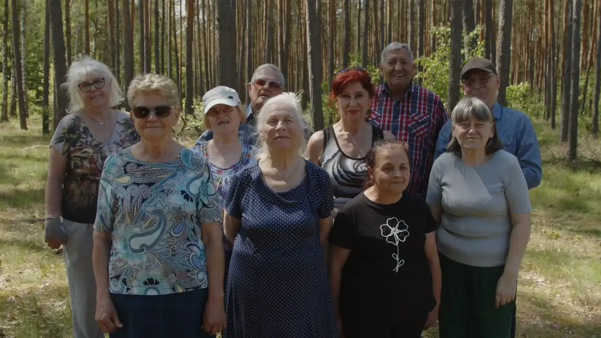 Na zdjęciu grupa starszych osób stojąca i pozująca do zdjęcia. Za nimi znajdują się drzewa.