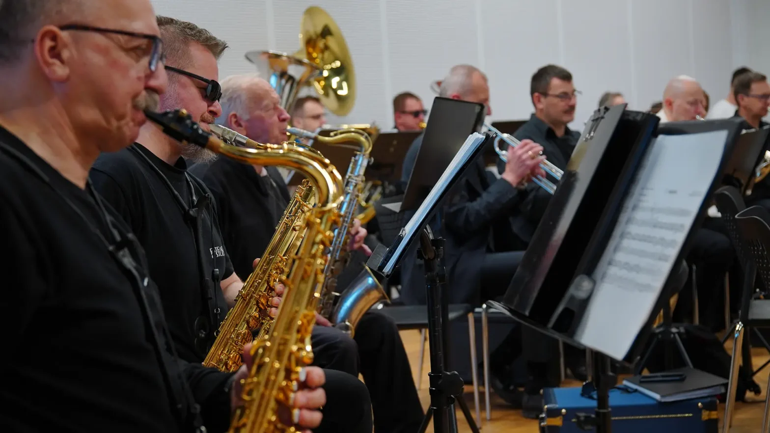 Na zdjęciu widzimy grupę mężczyzn grających na instrumentach. Na pierwszym planie trzech mężczyzn gra na saksofonach a przed nimi są stojaki z nutami.