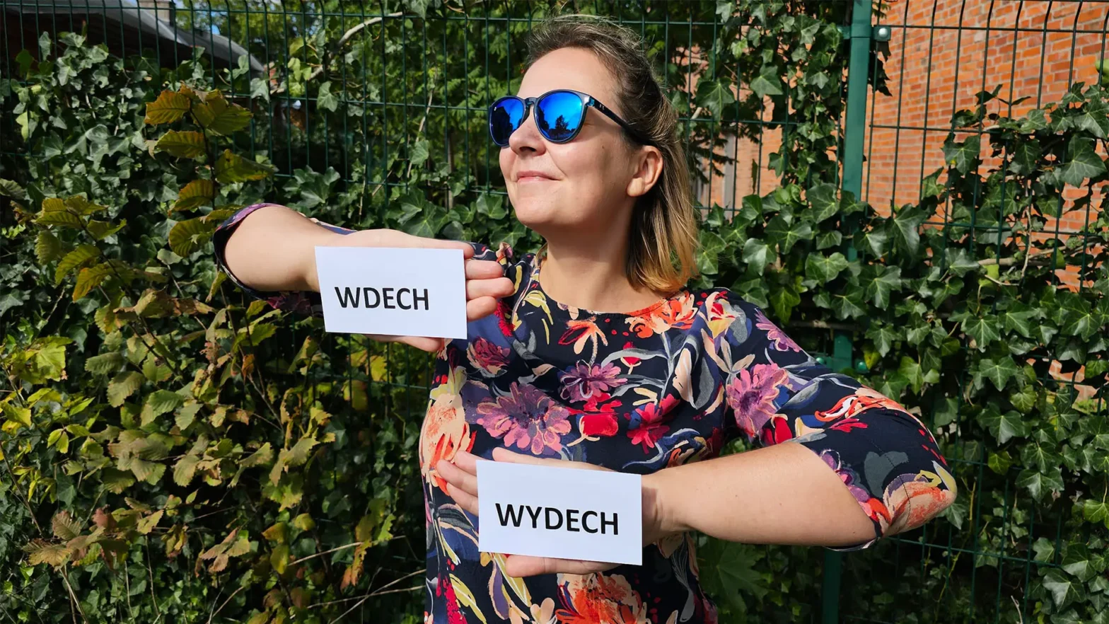 Na zdjęciu widać młoda kobietę ubraną w bluzkę w kolorowe kwiaty . Na na twarzy ma czarne okulary przeciwsłoneczne. Obie ręce ma zgięte w łokciach i skierowane do środka. Na prawym przedramieniu na doczepioną białą kartkę z czarnym, pogrubionym wyrazem WDECH. Na drugiej ręce skierowanej w prawą stronę ma karteczkę z napisem WYDECH. Postać ta stoi na tle ciemnozielonych krzewów, zza którego widać fragment ceglanego muru.