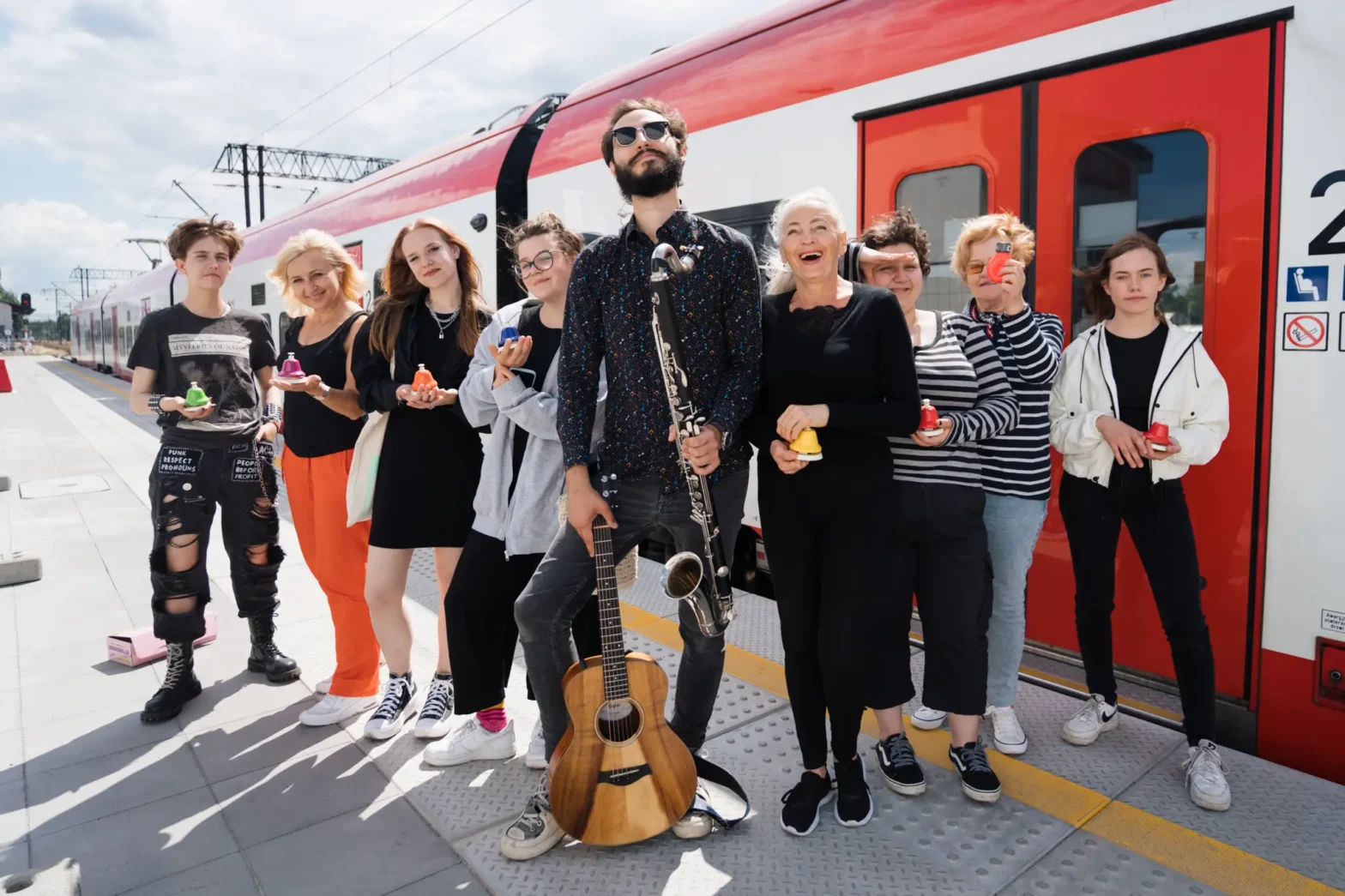 Grupa dziewięciu osób stojących na peronie przed czerwonobiałym pociągiem. Wśród nich osoby w różnym wieku i stylu, uśmiechnięte, trzymające w dłoniach kolorowe dzwonki ręczne. Na środku mężczyzna z brodą i okularami przeciwsłonecznymi, trzymający klarnet basowy. Mężczyzna oparty jest o gitarę akustyczną. Zdjęcie wykonane w słoneczny dzień, w radosnej i luźnej atmosferze. Zdjęcie zrobiła Monika Gut Jarecka