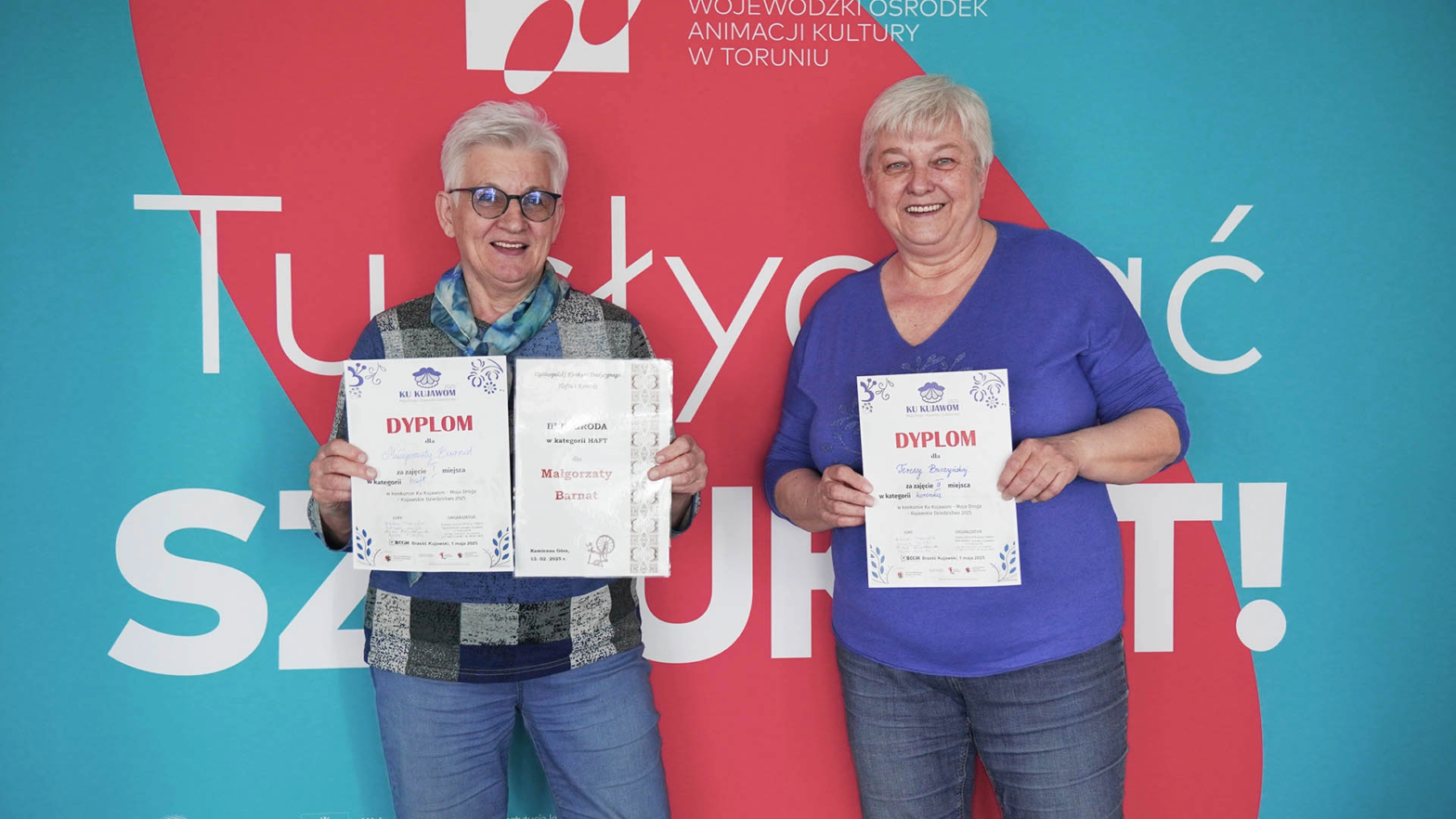 Na tle kolorowej ścianki z napisem Tu słychać SZTUKOT stoją Małgorzata Barnat i Genowefa Dzięgiel, trzymając w dłoniach zdobyte dyplomy i nagrody. Ich uśmiechy są szczere i spokojne – to moment radości z własnej pracy, z pasji do haftu i z docenienia w konkursie Ku Kujawom. Ich dłonie stworzyły coś pięknego, a teraz pokazują to światu z naturalną dumą.