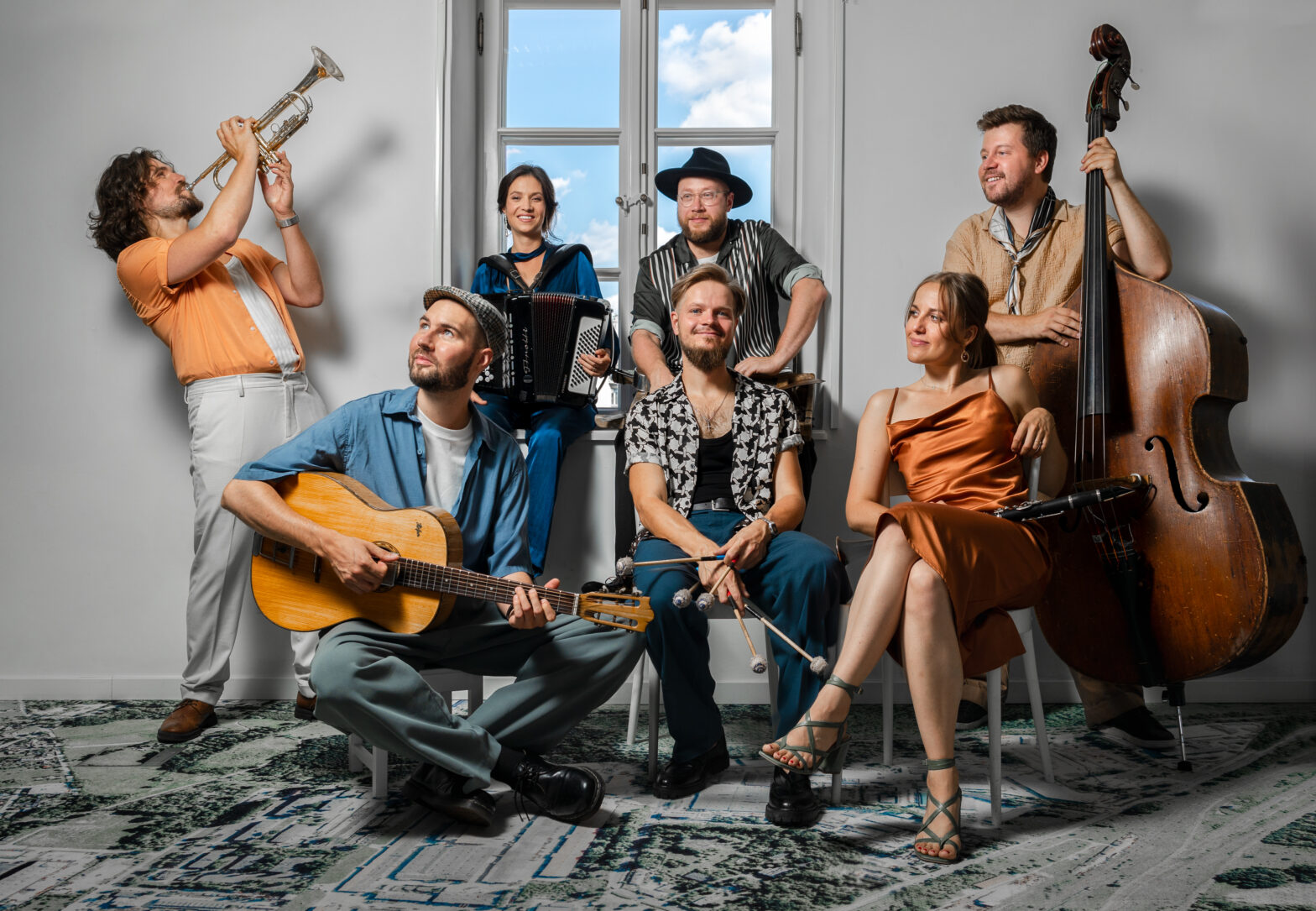 Portret zespołu muzycznego ukazuje siedem osób (kobiet i mężczyzn) z instrumentami: gitarą, kontrabasem, trąbką, skrzypcami i akordeonem. Pozują razem w jasnym pomieszczeniu z oknem w tle. Widać między nimi dobrą energię i swobodę.