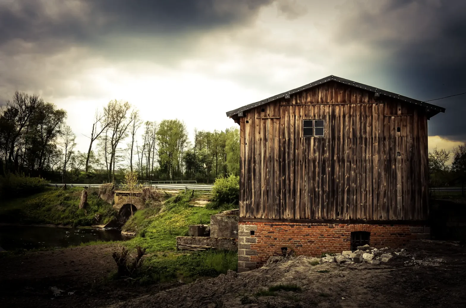 Drewniany budynek młyna z ceglanym fundamentem stojący nad rzeką, otoczony drzewami, pod pochmurnym niebem.