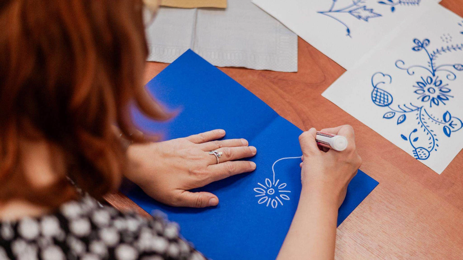 Kobieta w rudych włosach rysuje kwiatka białym pisakiem na niebieskiej kartce. Kobieta skierowana jest tyłem do kamery. Na stoliku widzimy również inne rysunki.
