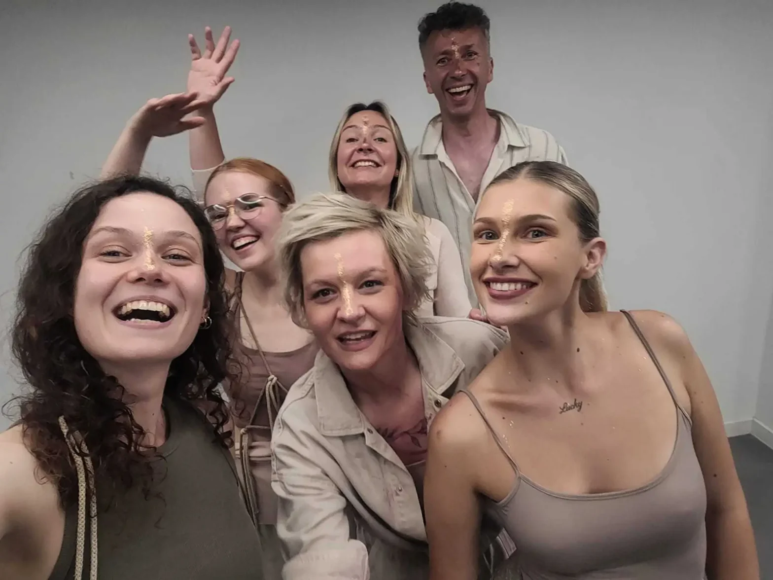 Grupa sześciu osób pozuje do zdjęcia w pomieszczeniu. Pięć kobiet i jeden mężczyzna uśmiechają się do aparatu, dwie kobiety machają rękami. Na ich twarzach widać radość i entuzjazm.