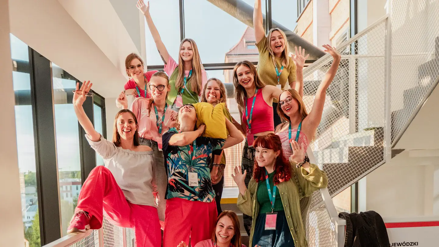 Na zdjęciu grupa wesoło uśmiechjących się kobiet, które także machaja do zdjęcia. Ubrane są w kolorwe ubrania,