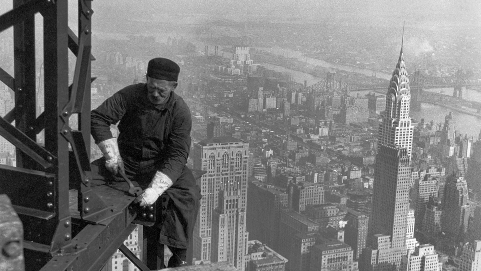 Czarnobiała fotografia wykonana z dużej wysokości. Na pierwszym planie, po prawej stronie, widać mężczyznę siedzącego na metalowym rusztowaniu. Jest ubrany w robocze spodnie i koszulę z długim rękawem. W jednej ręce trzyma narzędzie i dokręca element konstrukcji. W tle rozciąga się panorama miasta: dachy budynków i ulice, które wydają się bardzo małe z tej perspektywy. Zdjęcie oddaje zarówno odwagę, jak i codzienność pracy robotników budowlanych na początku dwudziestego wieku.