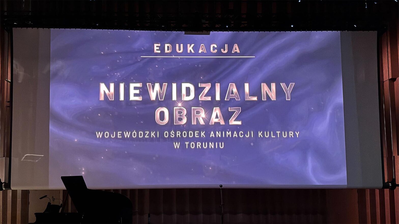 Na zdjęciu widać ekran, na którym wyświetlony jest złoty napis na niebieskim tle: Edukacja. Niewidzialny Obraz. Wojewódzki Ośrodek Animacji Kultury w Toruniu.