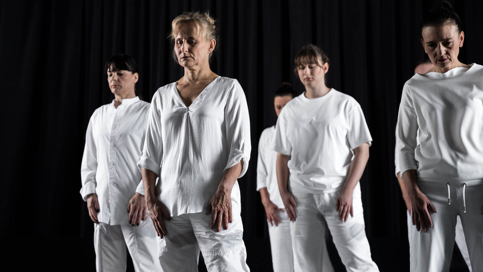 Grupa kobiet stoi w półkolu na scenie, w czarnym tle. Wszystkie mają na sobie białe stroje: koszule, bluzy i spodnie. Stoją nieruchomo, z opuszczonymi rękami i głowami lekko pochylonymi w dół.