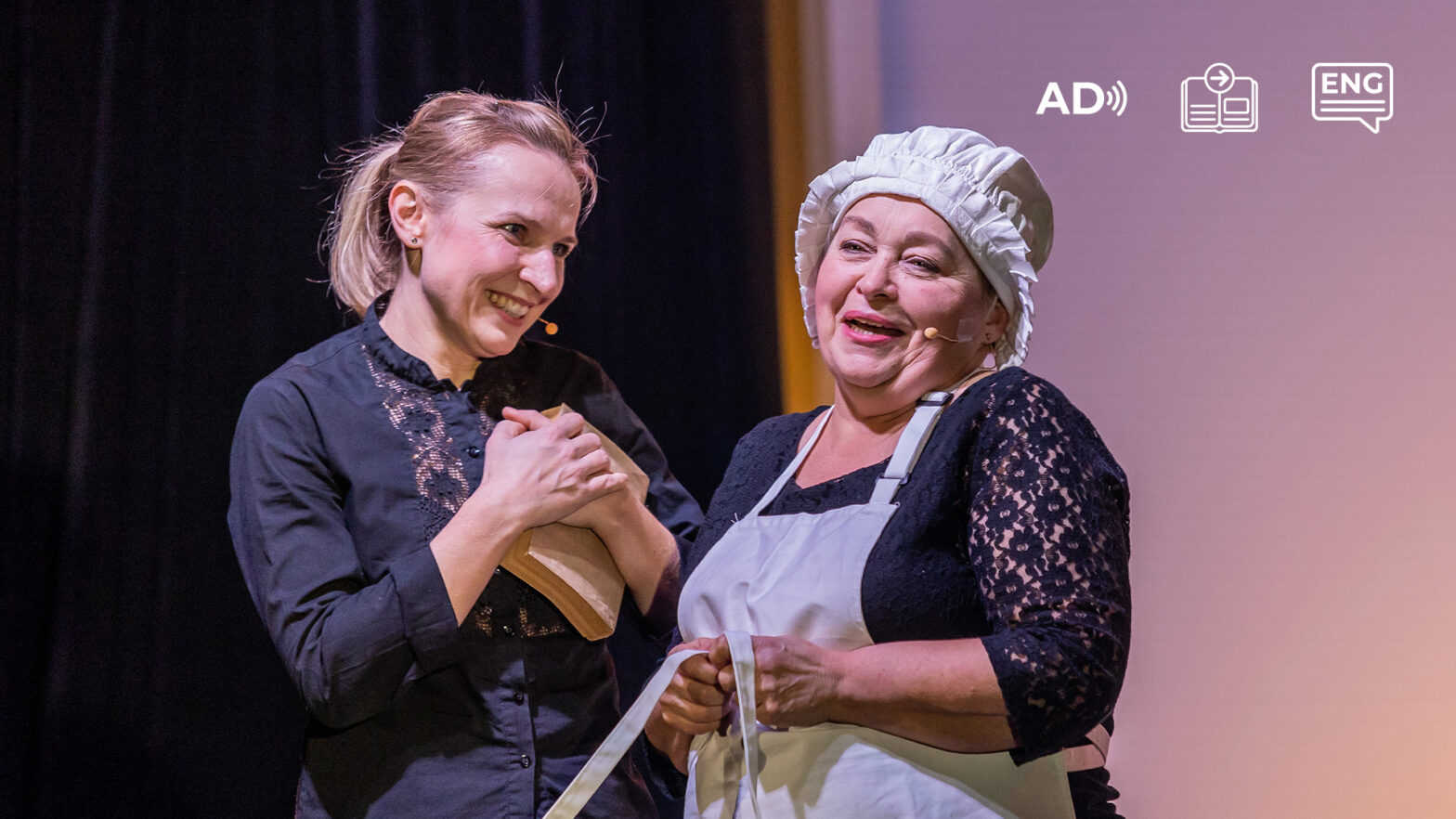 Na zdjęciu widać dwie aktorki ze spektaklu Budowniczki. Stoją blisko siebie w centrum kadru. Jedna z nich przytula książkę do piersi i uśmiecha się do aktorki stojącej po prawej stronie. Druga aktorka patrzy przed siebie z uśmiechem. Ma na sobie biały kucharski czepek i fartuch, pod spodem czarną bluzkę. W prawym górnym rogu znajduje się piktogram audiodeskrypcji, przewodnika i napisów w języku angielskim.