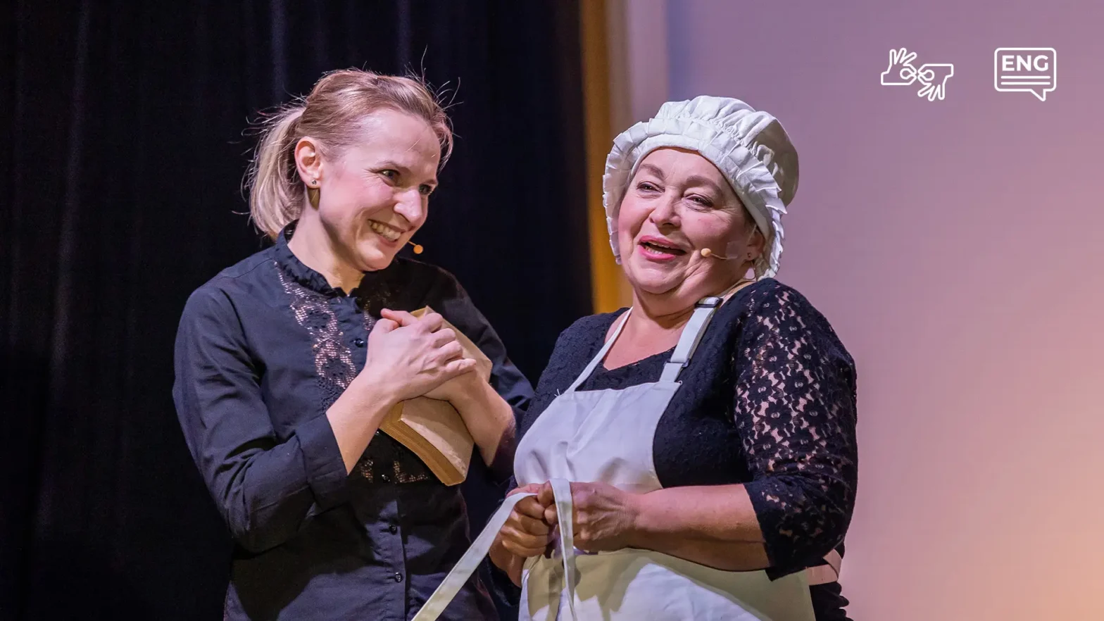 Na zdjęciu dwie aktorki grające w spektaklu Budowniczki. Stoją w centrum zdjęcia blisko siebie. Jedna z nim trzyma w rękach książke i prztula ją do piersi, uśmiechając się do drugiej aktorki po prawekj stronie. Druga aktorska z uśmiechem patrzy prtzed siebie. Ubrana jest w biały  kucharski czepek i biały fartuch. pod spodem ma czarną bluzkę.