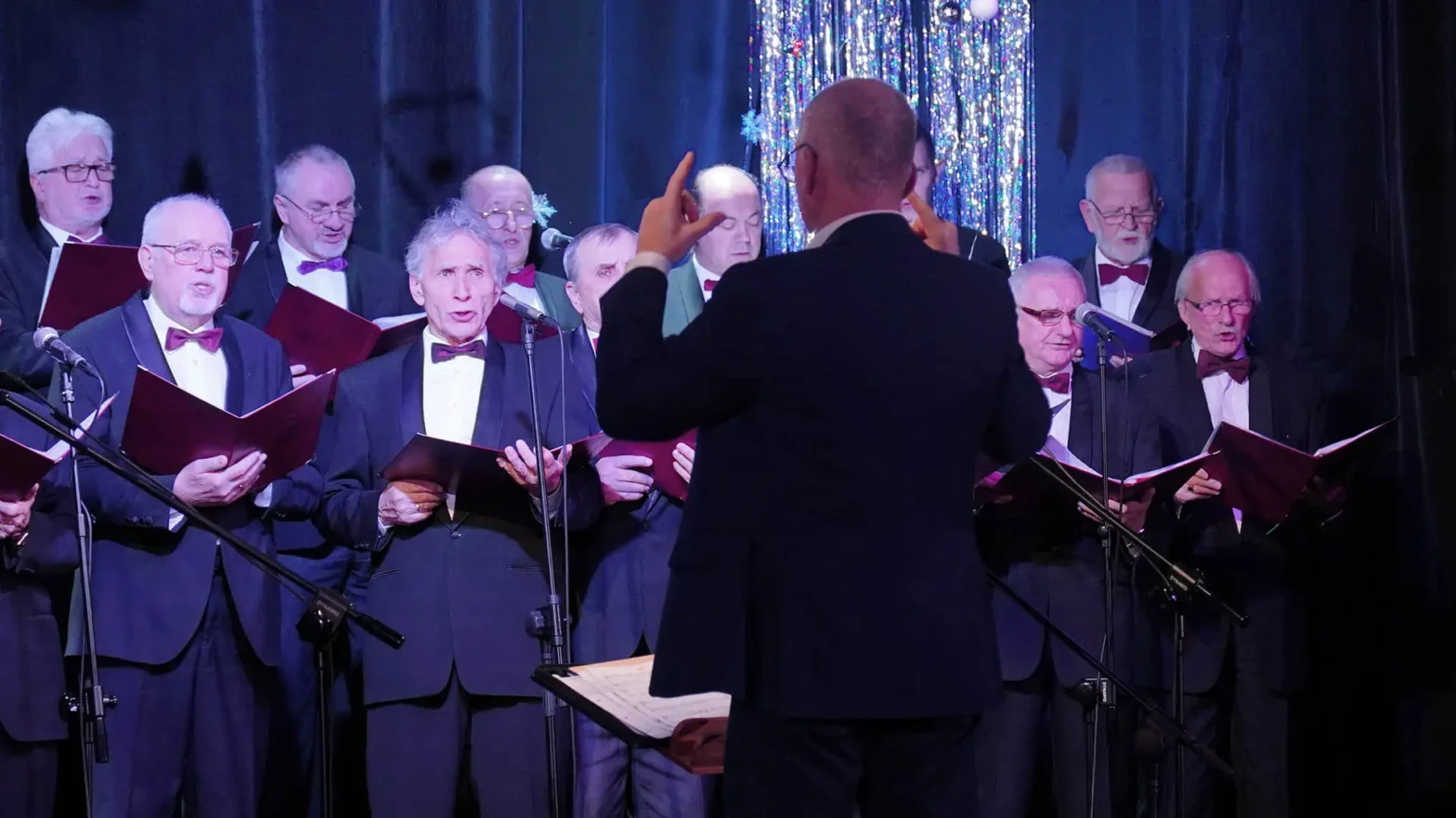 Na zdjęciu Chór Męski Kopernikus. W centralnym punkcie zdjęcia znajduje się dyrygent, stojący tyłem do aparatu. Przed nim stoi grupa mężczyzn trzymają w ręku zeszyty z piosenkami i którzy śpiewają do mikrofonów stojących przed nimi.