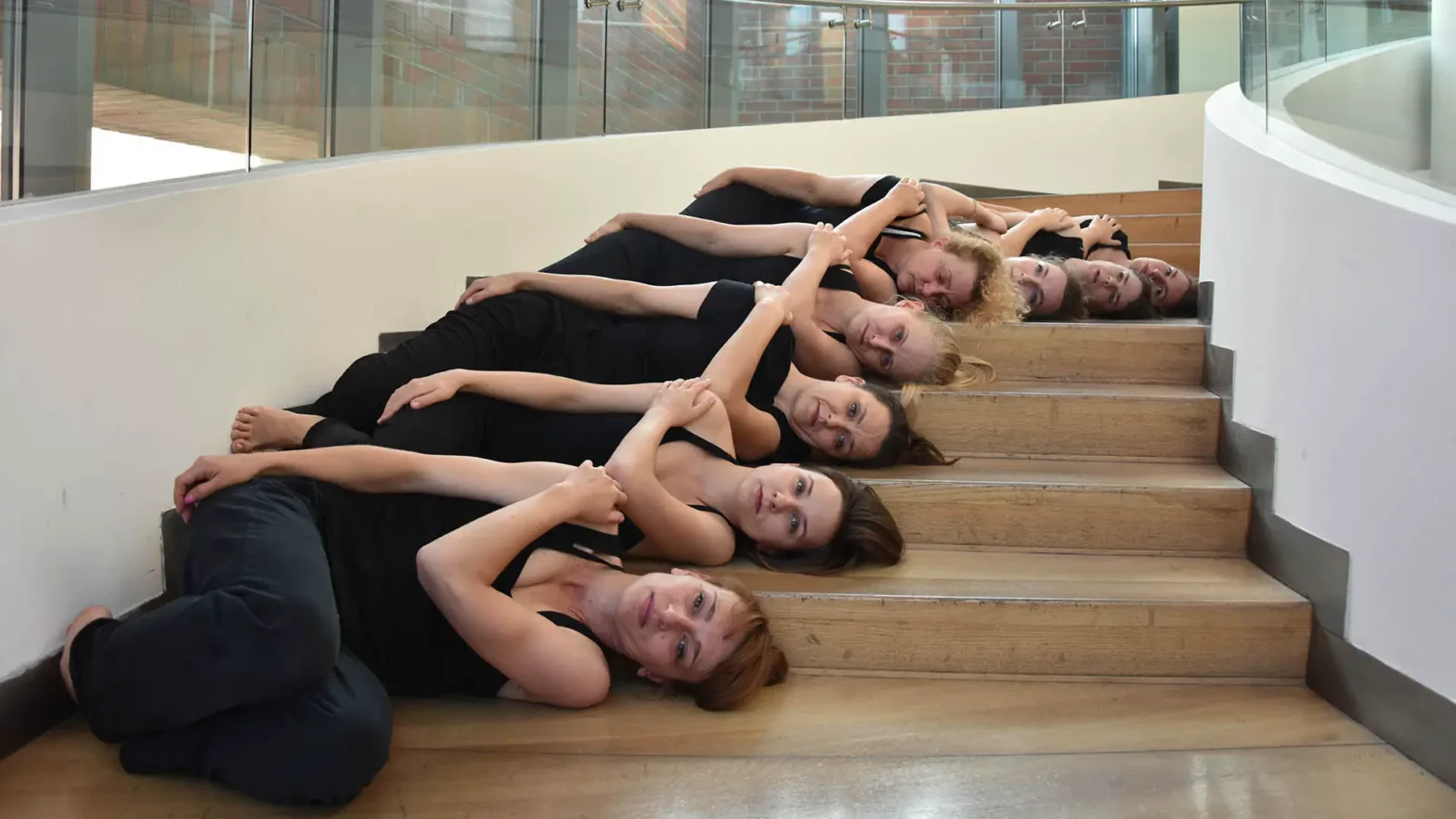 Grupa kobiet w czarnych strojach leży na schodach wewnątrz budynku. Ułożone są w jednej linii, głowami opartymi o stopnie, jedna za drugą. Ich ciała tworzą falującą kompozycję biegnącą wzdłuż schodów.