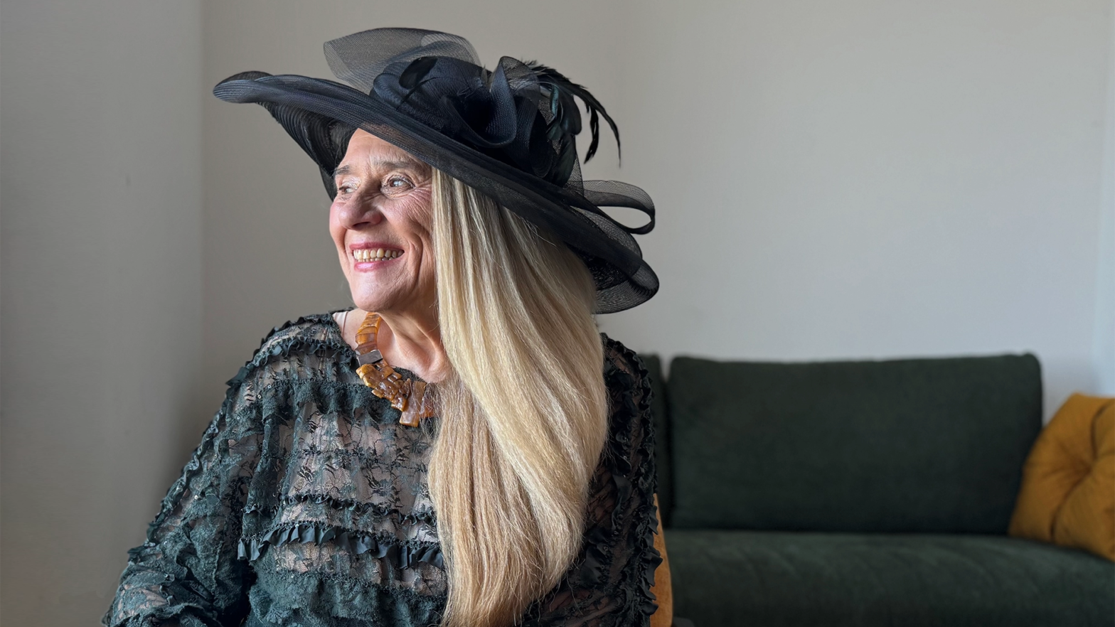 Na zdjęciu starsza blond kobieta, uśmiechająca się i patrząca w lewą stronę. Na głowie ma piękny czarny elegancki kapelusz z czarną woalką i piórem. Ubrana jest w czarną bluzkę z delikatnymi falbankami, a na szyi ma brązowy naszyjnik z kamieniami. Za nią w tle znajduję się zielona kanapa z żółtą poduszką.