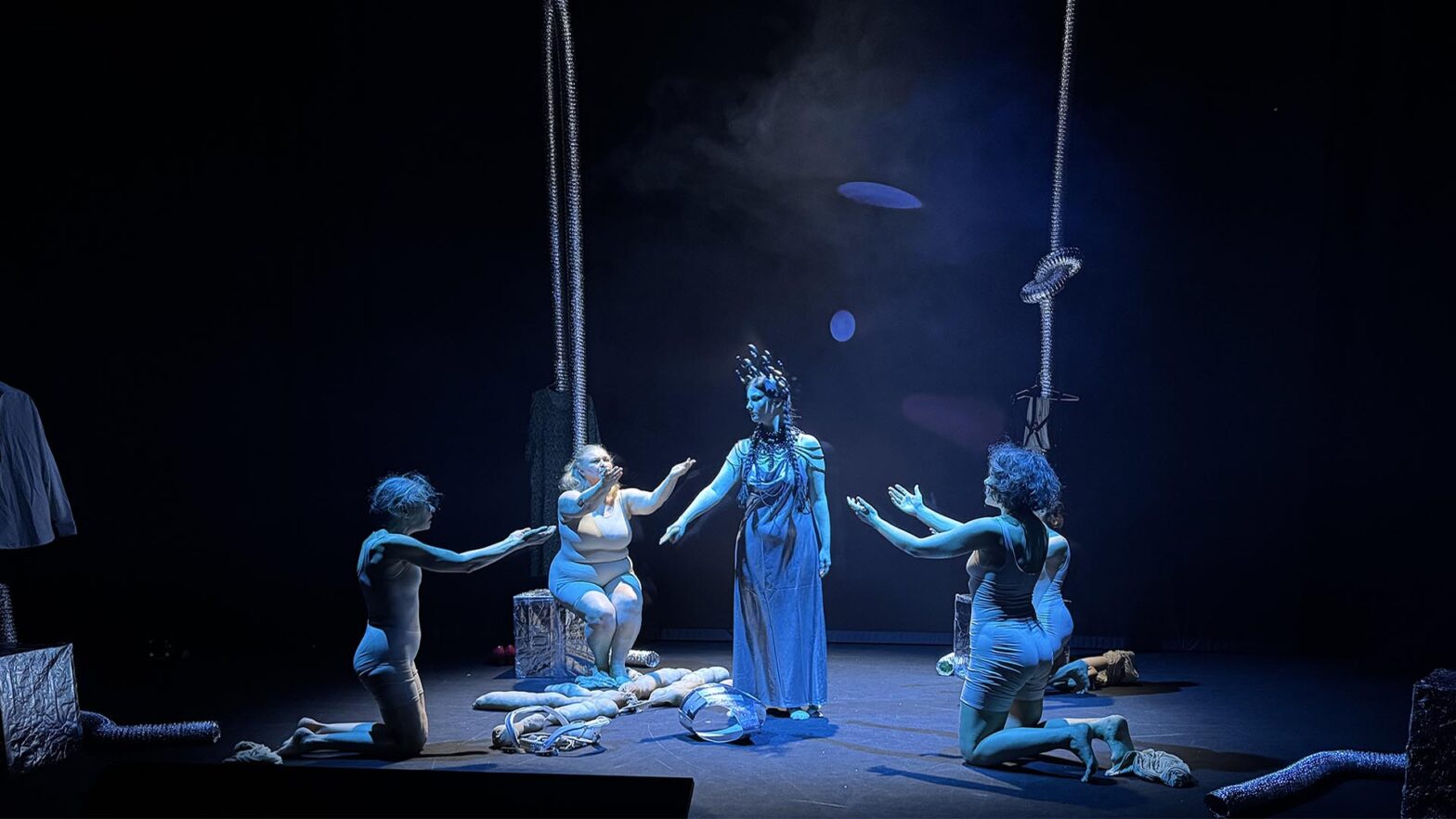 Scena teatralna oświetlona chłodnym, niebieskim światłem. W centrum stoi kobieta w długiej sukni. Wokół niej cztery kobiety klęczą i wyciągają ręce w jej stronę. Nad sceną wiszą grube srebrne rury. Na podłodze leżą przedmioty i materiały. Całość ma skupiony, trochę rytualny nastrój.