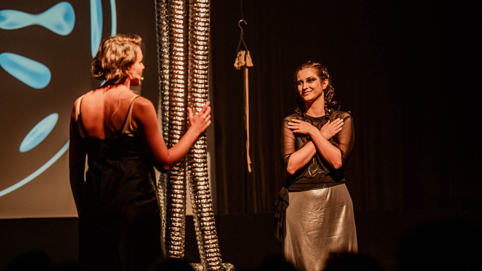 Dwie aktorki występują na scenie podczas spektaklu. Postać po prawej stronie ma zaplecione warkocze, ciemny makijaż i srebrną spódnicę. Trzyma dłonie skrzyżowane na piersiach i uśmiecha się do drugiej kobiety, która stoi tyłem do obiektywu w czarnej sukni. Między nimi zwisają ze stropu srebrne, karbowane rury, a w tle widać ekran z niebieską grafiką.