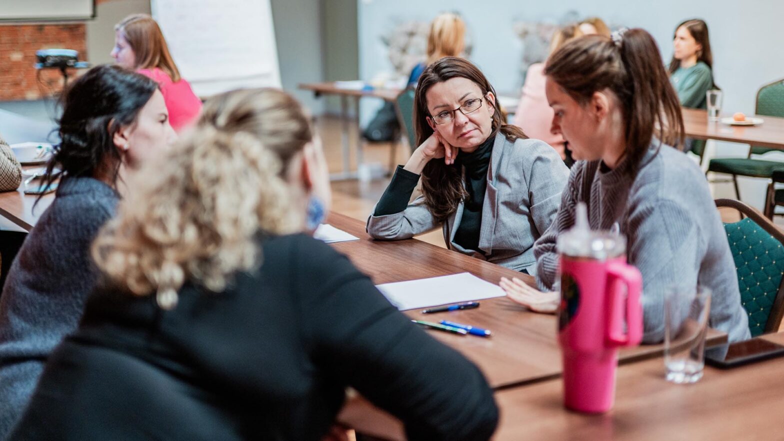 Grupa kobiet siedzi przy podłużnym, drewnianym stole w jasnym pomieszczeniu podczas spotkania. Na pierwszym planie kobieta w okularach i szarym żakiecie z uwagą słucha swojej rozmówczyni, opierając głowę na dłoni. W tle widać inne osoby siedzące przy stolikach.