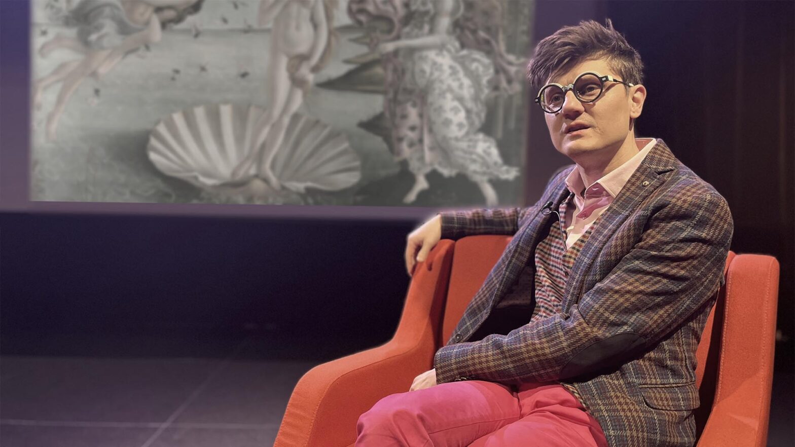 Na zdjęciu Łukasz Wudarski. Mężczyzna w okularach o grubych, okrągłych oprawkach siedzi w czerwonym fotelu na tle dużego ekranu. Ma na sobie kraciastą marynarkę, koszulę w paski i jaskraworóżowe spodnie. Patrzy w górę z zaciekawieniem. Na ekranie za nim wyświetlany jest fragment klasycznego obrazu pod tytułem Narodziny Wenus autorstwa Sandra Boticzellego.