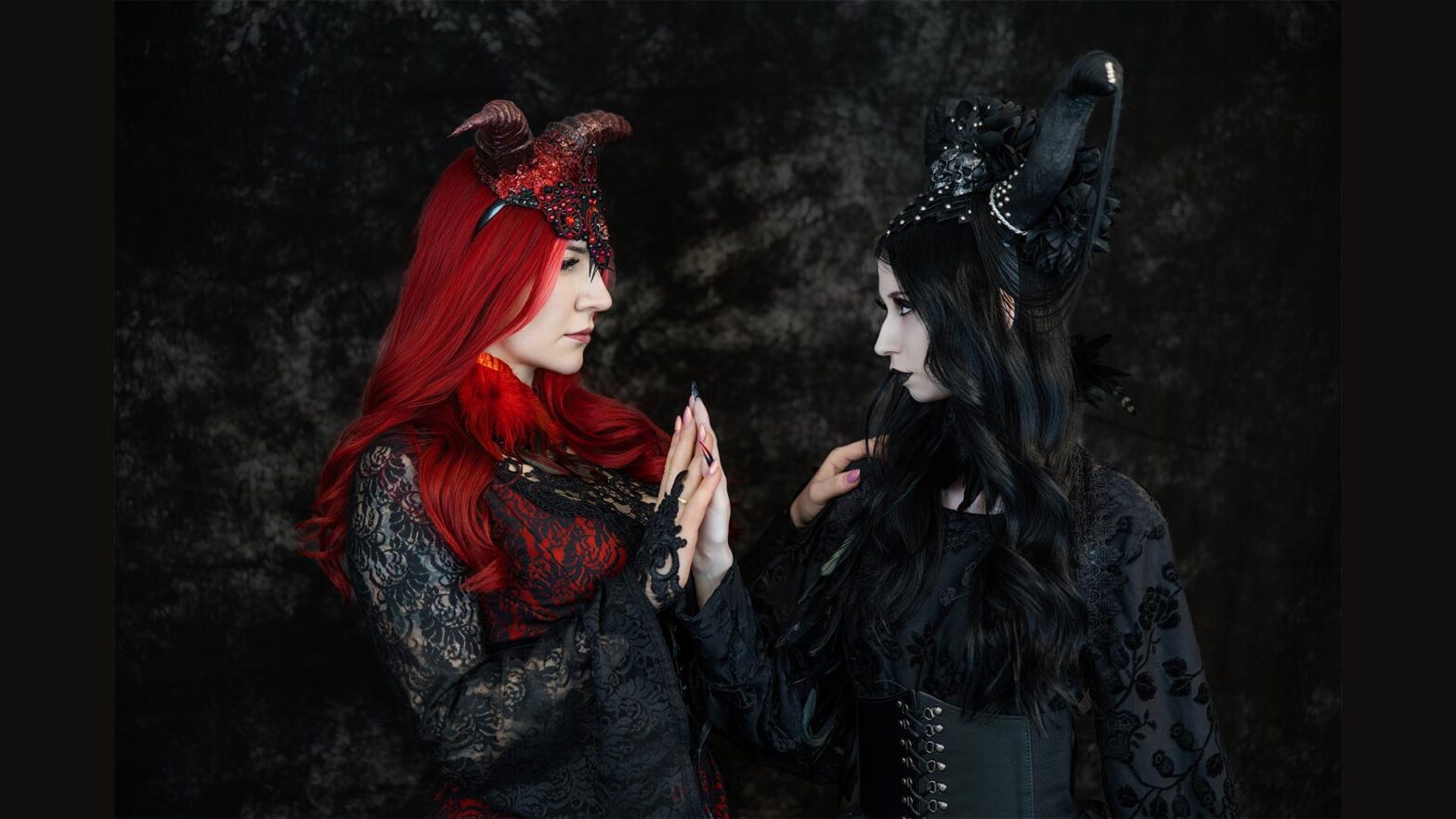 Dwie kobiety w stylizowanych kostiumach fantazy stoją naprzeciwko siebie na ciemnym tle. Postać po lewej ma długie, intensywnie czerwone włosy, czerwone rogi i koronkową suknię. Postać po prawej ma czarne włosy, ciemny makijaż, czarny strój oraz wysokie, zdobione rogi. Kobiety stykają się dłońmi i patrzą sobie prosto w oczy.