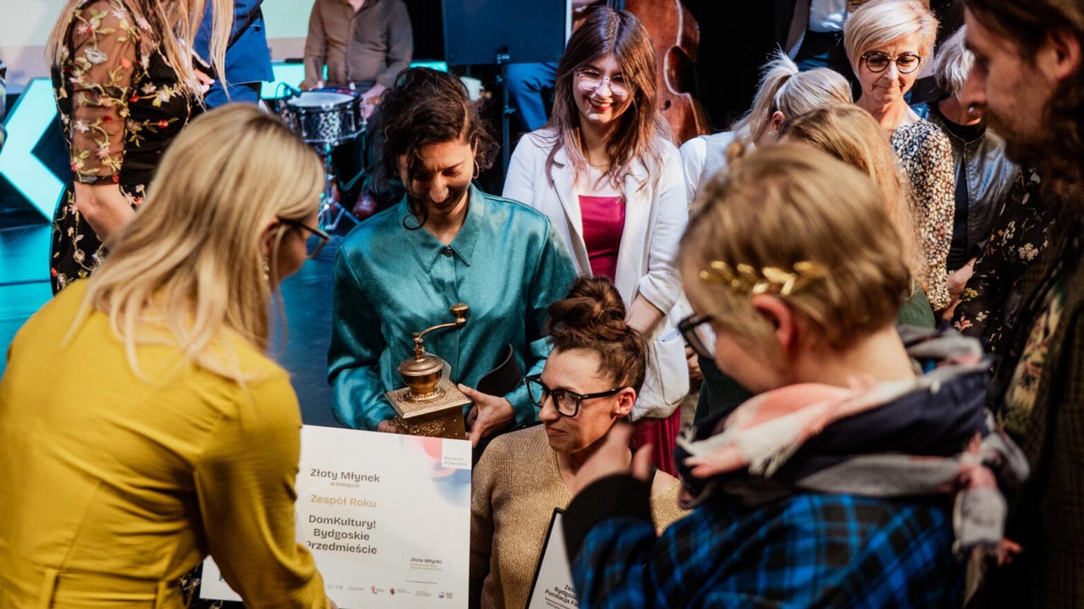 Zdjęcie przedstawia grupę osób zgromadzonych wokół kobiety siedzącej na pierwszym planie podczas wręczenia nagrody. Jedna z osób przekazuje dyplom, a inna trzyma statuetkę przypominającą młynek. Wszyscy są skupieni na tej chwili. Na dyplomie widoczny jest napis Złoty Młynek oraz informacja o nagrodzie dla zespołu.