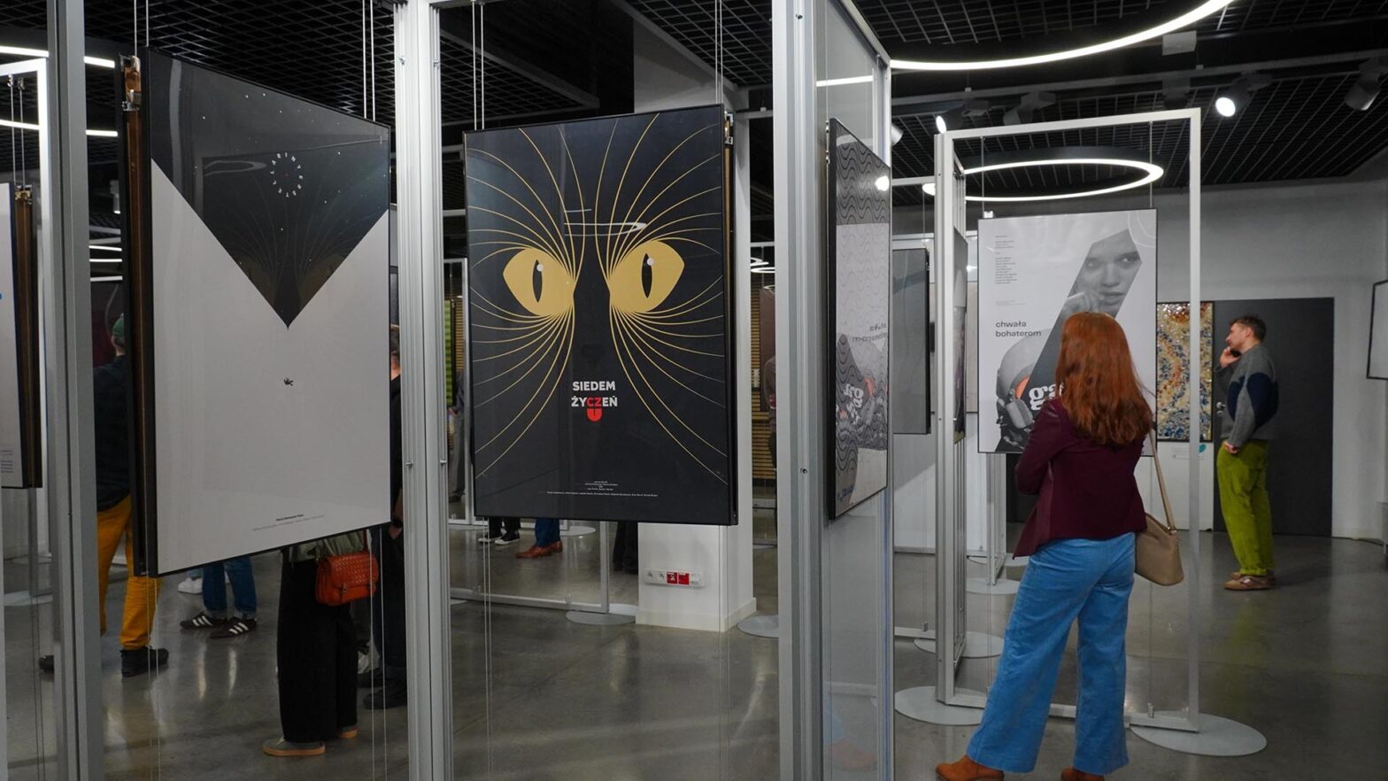 Parter Młynów Kultury z wystawą plakatów filmowych autorstwa Mirka Gurzyńskiego. Ekspozycja składa się z prac zawieszonych na linkach w metalowych stelażach. Na pierwszym planie widać plakat z dwiema dużymi, żółtymi kocimi oczami na czarnym tle i napisem Siedem życzeń. W tle znajdują się inne plakaty oraz zwiedzający, w tym kobieta z rudymi włosami w niebieskich spodniach, która tyłem do obiektywu ogląda wystawę.