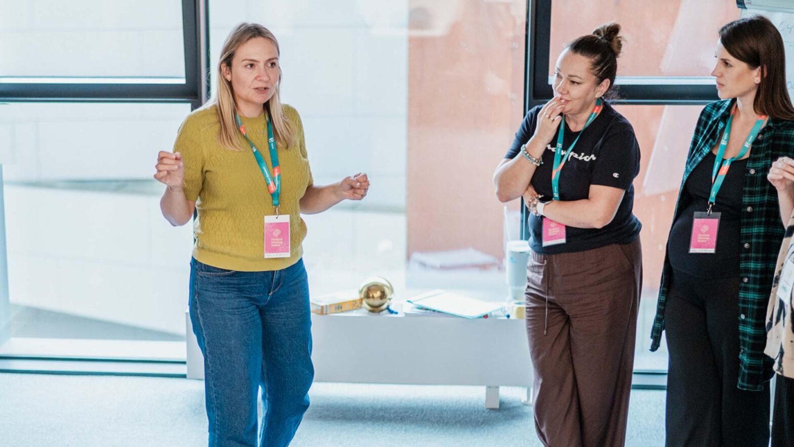 Kobieta stoi i mówi do grupy. Ma na sobie żółty sweter i identyfikator na szyi. Obok niej stoją dwie inne kobiety i słuchają. W tle duże okna i jasne wnętrze. Atmosfera skupienia i wspólnej pracy.