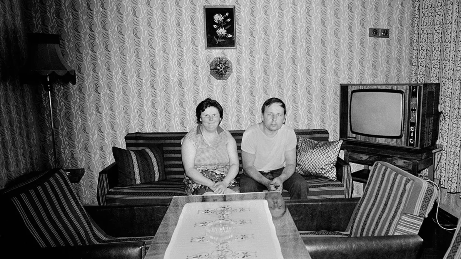 Czarnobiała fotografia przedstawiająca parę siedzącą na kanapie w pokoju urządzonym w stylu z drugiej połowy dwudziestego wieku. Ściany są pokryte wzorzystą tapetą w pionowe pasy z motywem roślinnym. Po prawej stronie widoczny jest stary telewizor kineskopowy w drewnianej obudowie. Przed kanapą stoi niski stolik z ozdobną serwetą.