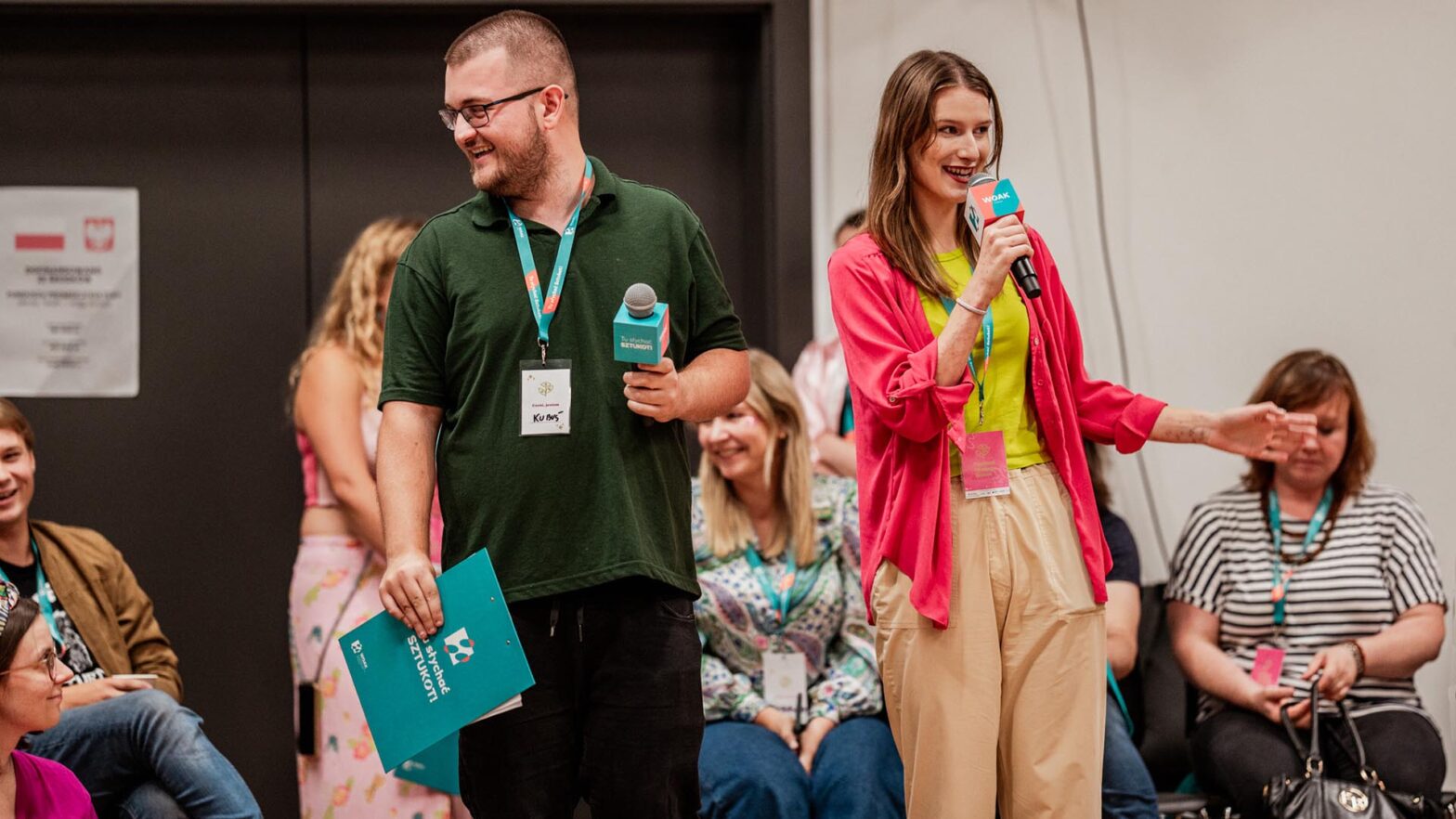 Zdjęcie przedstawia kobietę i mężczyznę prowadzących wydarzenie. Stoją obok siebie i trzymają mikrofony, zwracając się do publiczności. Kobieta mówi do mikrofonu, gestykulując ręką, a mężczyzna uśmiecha się, trzymając materiały w dłoni. W tle widoczne są siedzące osoby uczestniczące w spotkaniu.