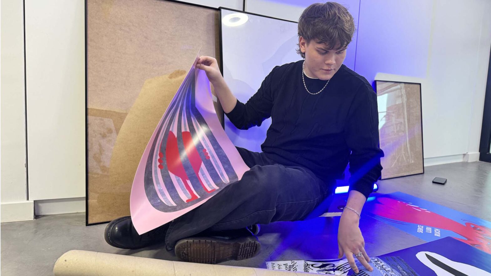 Zdjęcie przedstawia osobę siedzącą na podłodze we wnętrzu, przeglądającą i układającą plakaty. Trzyma w rękach jeden z nich, a obok leżą kolejne wydruki. W tle widoczne są oprawione grafiki oparte o ścianę. Scena ma spokojny charakter.