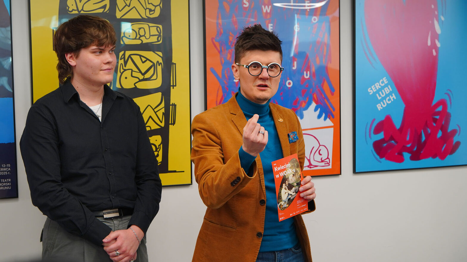 Zdjęcie przedstawia dwie osoby stojące we wnętrzu przed ścianą z kolorowymi plakatami. Jedna z nich przemawia i gestykuluje ręką, trzymając w drugiej ręce materiały informacyjne. Druga osoba stoi obok i słucha. W tle widoczne są plakaty z graficznymi ilustracjami w intensywnych kolorach.